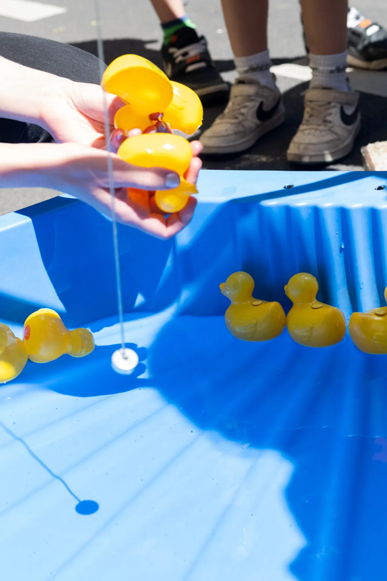 Eine Person angelt gelbe Plastikenten in einem blauen Becken, wahrend Kinderfusse im Hintergrund zu sehen sind.