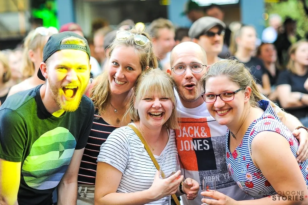 Eine Gruppe gut gelaunter, lachender Menschen posiert fur ein Foto auf einer belebten Veranstaltung.