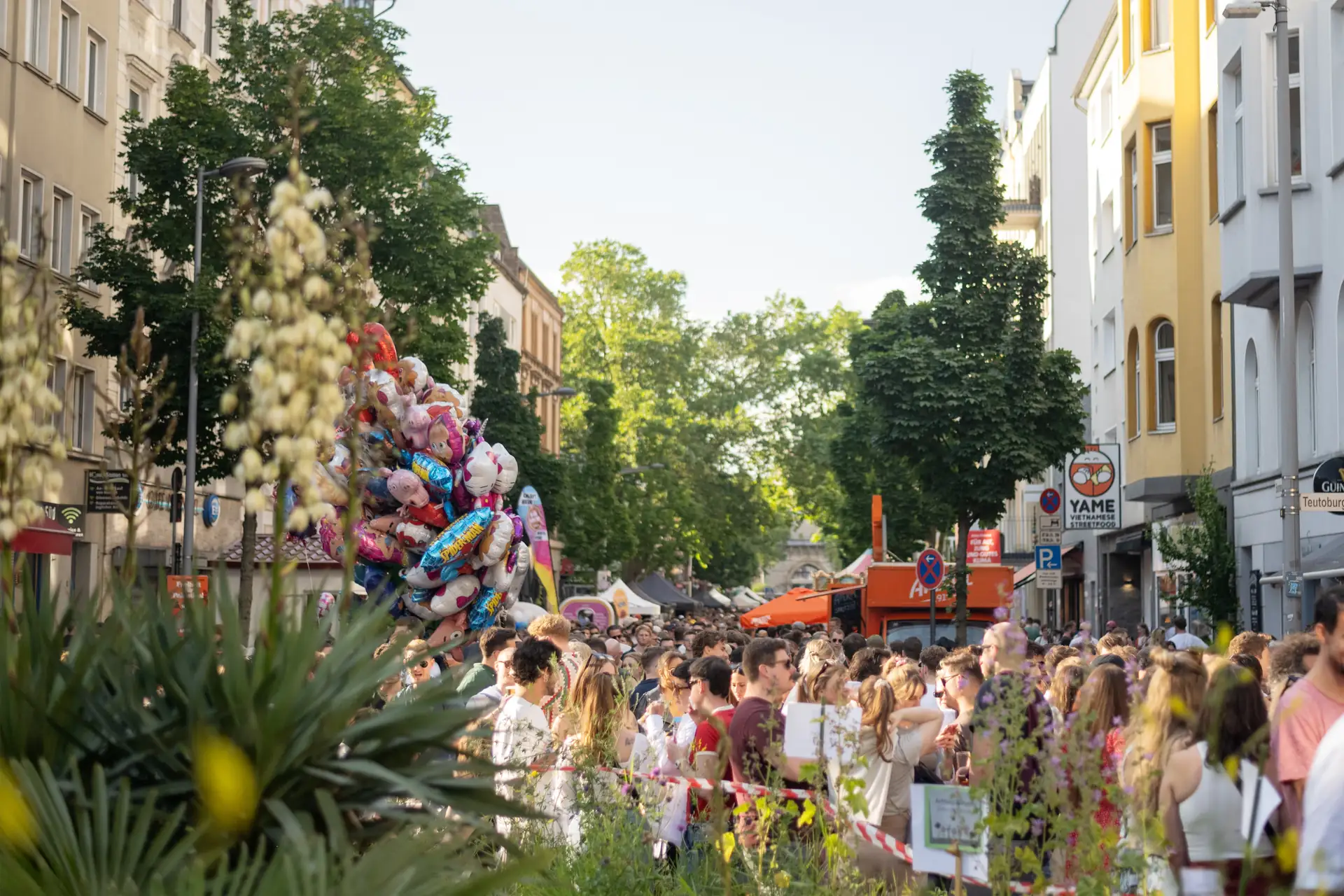 Eine grosse Menschenmenge drangt sich an einem sonnigen Tag auf einer Strasse, viele halten bunte Luftballons.