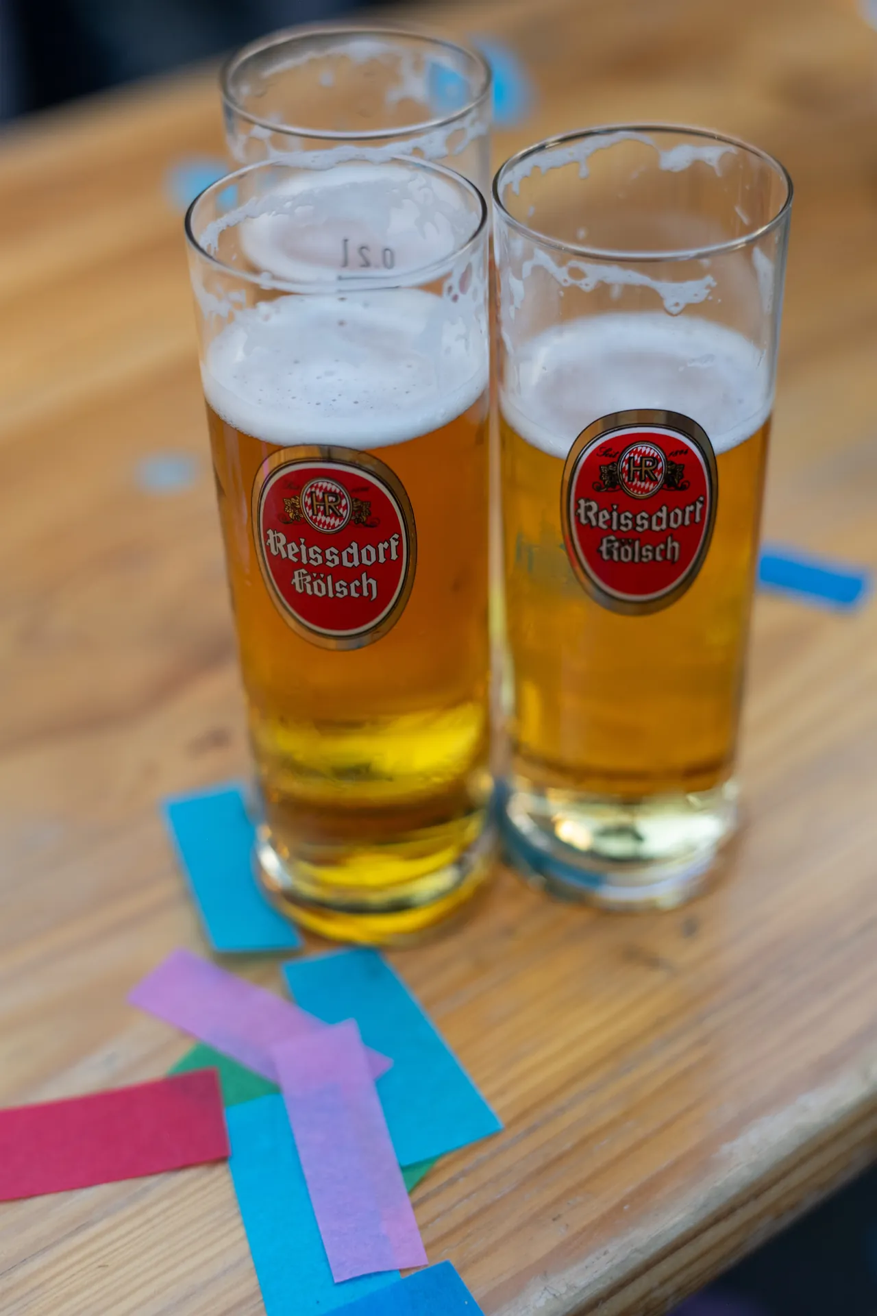 Drei fast volle Kolschglaser der Marke Reissdorf stehen auf einem Tisch, bunte Papierschnipsel liegen daneben.