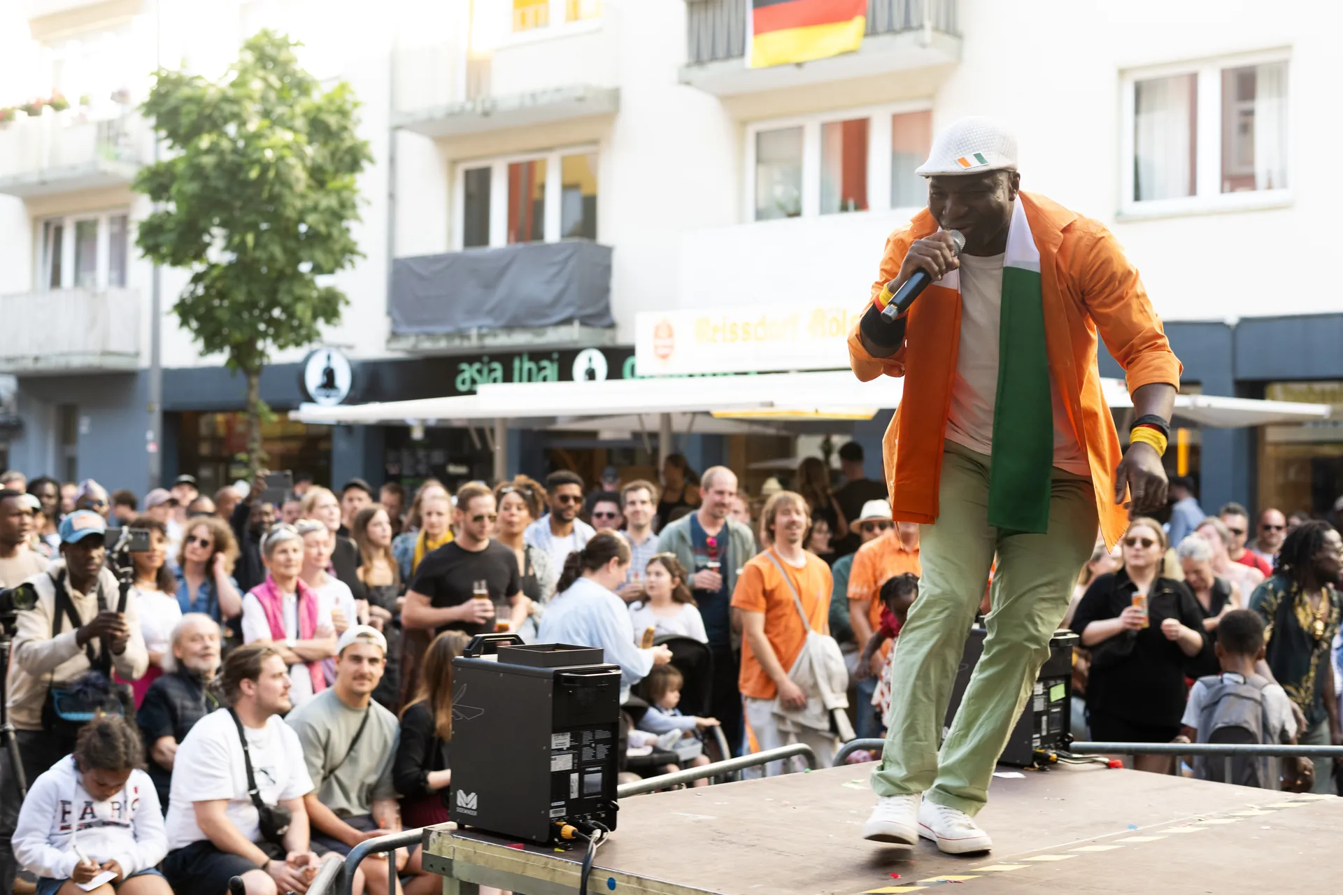 Ein dunkelhautiger Sanger in orangefarbener Jacke und Mutze performt auf einer Buhne vor Publikum.